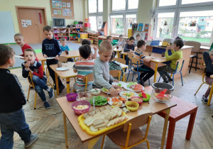 Dzieci siedzą przy stolikach, jedzą śniadanie, na głównym planie szwedzki stół z potrawami